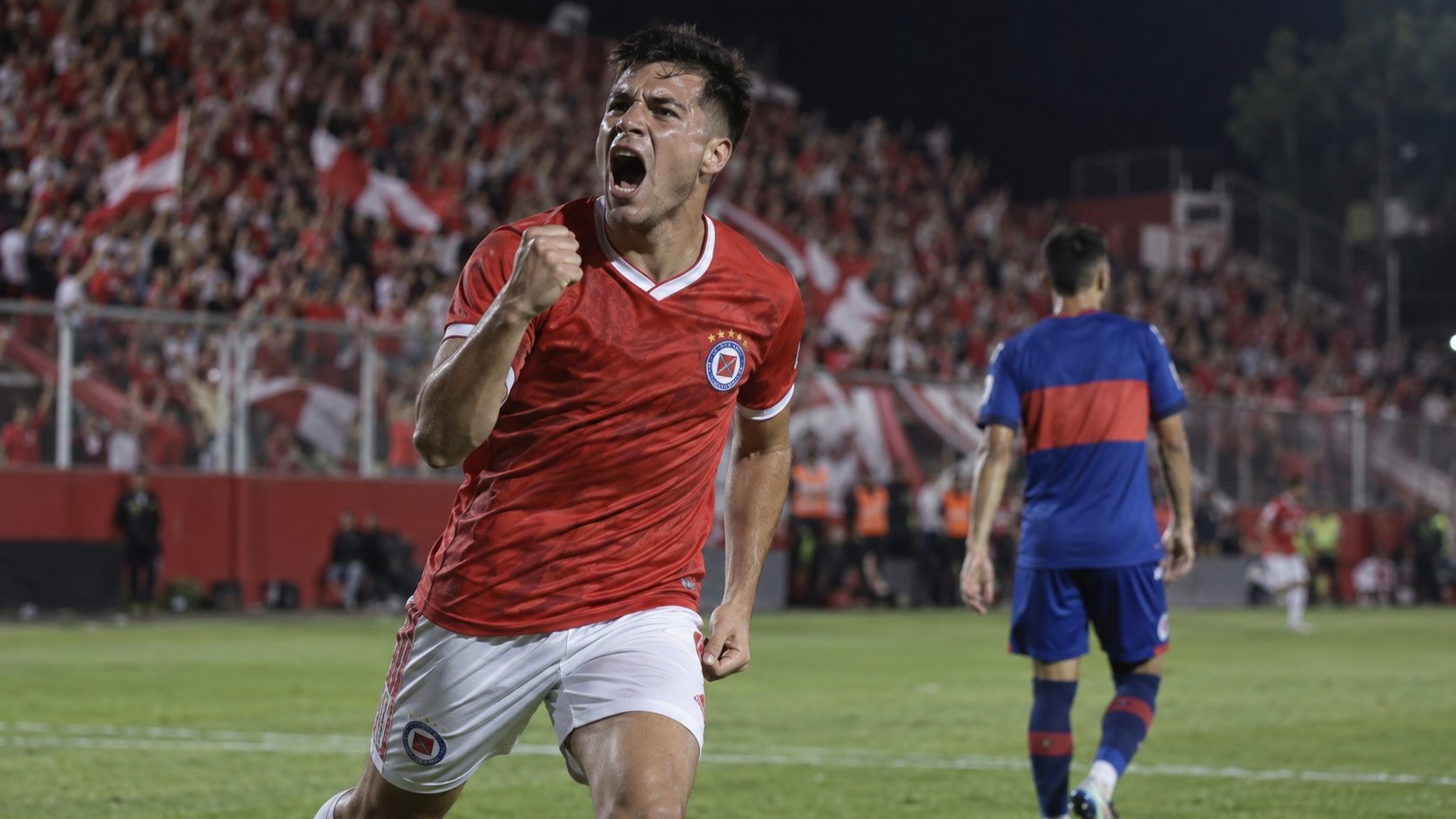 Ilustración editorial generada con IA: de noche, en un estadio con las tribunas llenas de hinchas de rojo, un futbolista con la camiseta roja de Argentinos Juniors festeja un gol con el puño cerrado mientras, al fondo y desenfocado, un rival con camiseta azul con franja roja queda de espaldas; no es una fotografía del partido ni representa a jugadores reales.