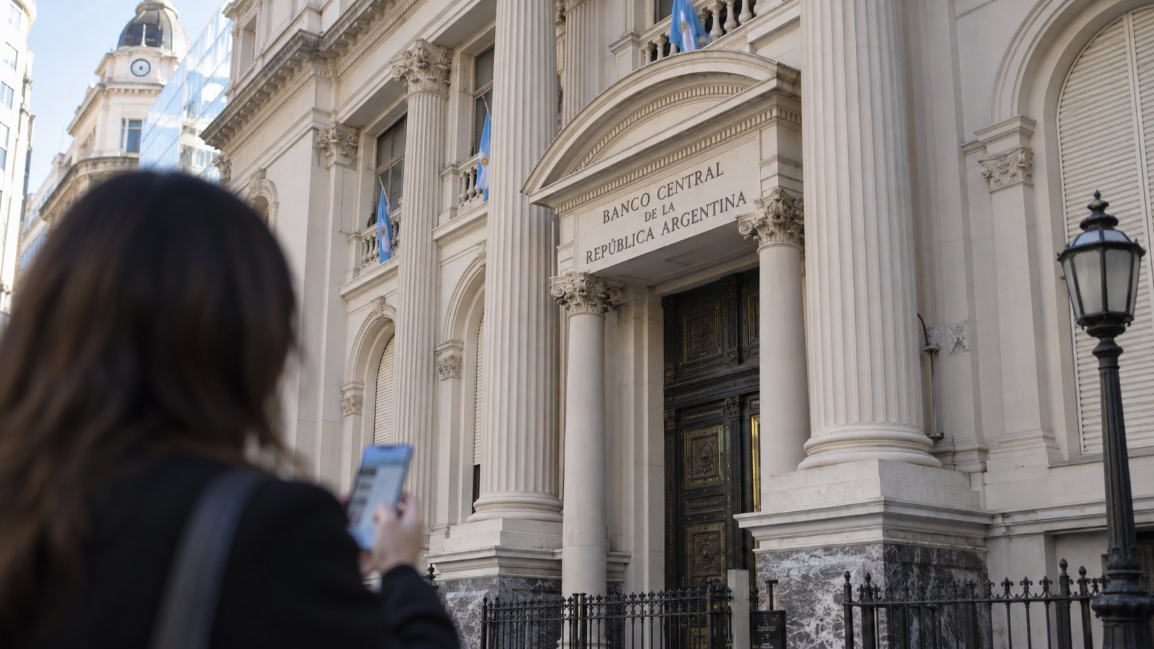 Ilustración editorial generada con IA: la fachada histórica del Banco Central de la República Argentina en el microcentro porteño, con su nombre sobre la entrada y banderas argentinas, y en primer plano una mujer de espaldas que mira su celular; composición conceptual, no es una fotografía del anuncio.