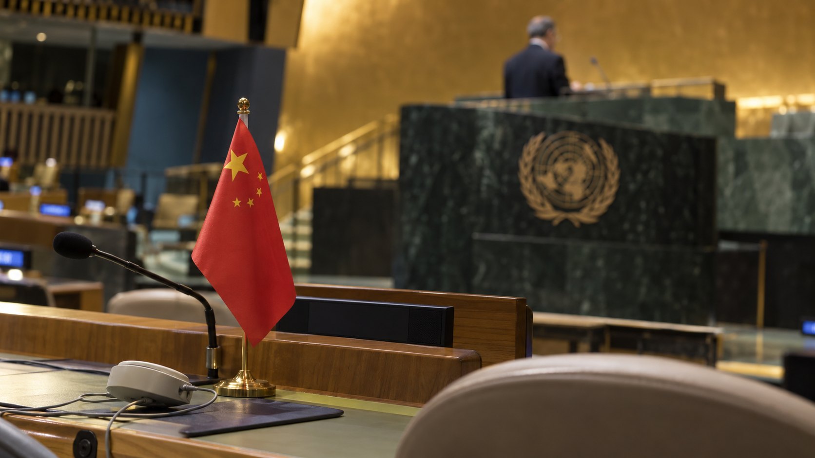 Ilustración editorial generada con IA: bandera de mesa de la República Popular China junto a un micrófono en una banca de delegados del salón de la Asamblea General de la ONU, con el podio de mármol verde y el emblema de las Naciones Unidas al fondo; no es una fotografía del discurso.