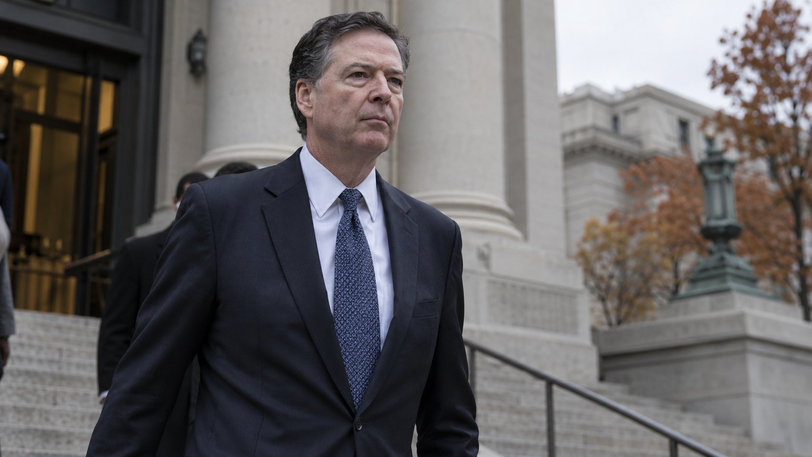 Ilustración editorial generada con IA: James Comey, ex director del FBI, de traje oscuro, baja serio la escalinata de un tribunal federal en un día nublado de otoño; composición representativa, no es una fotografía del lunes ni de una comparecencia real.