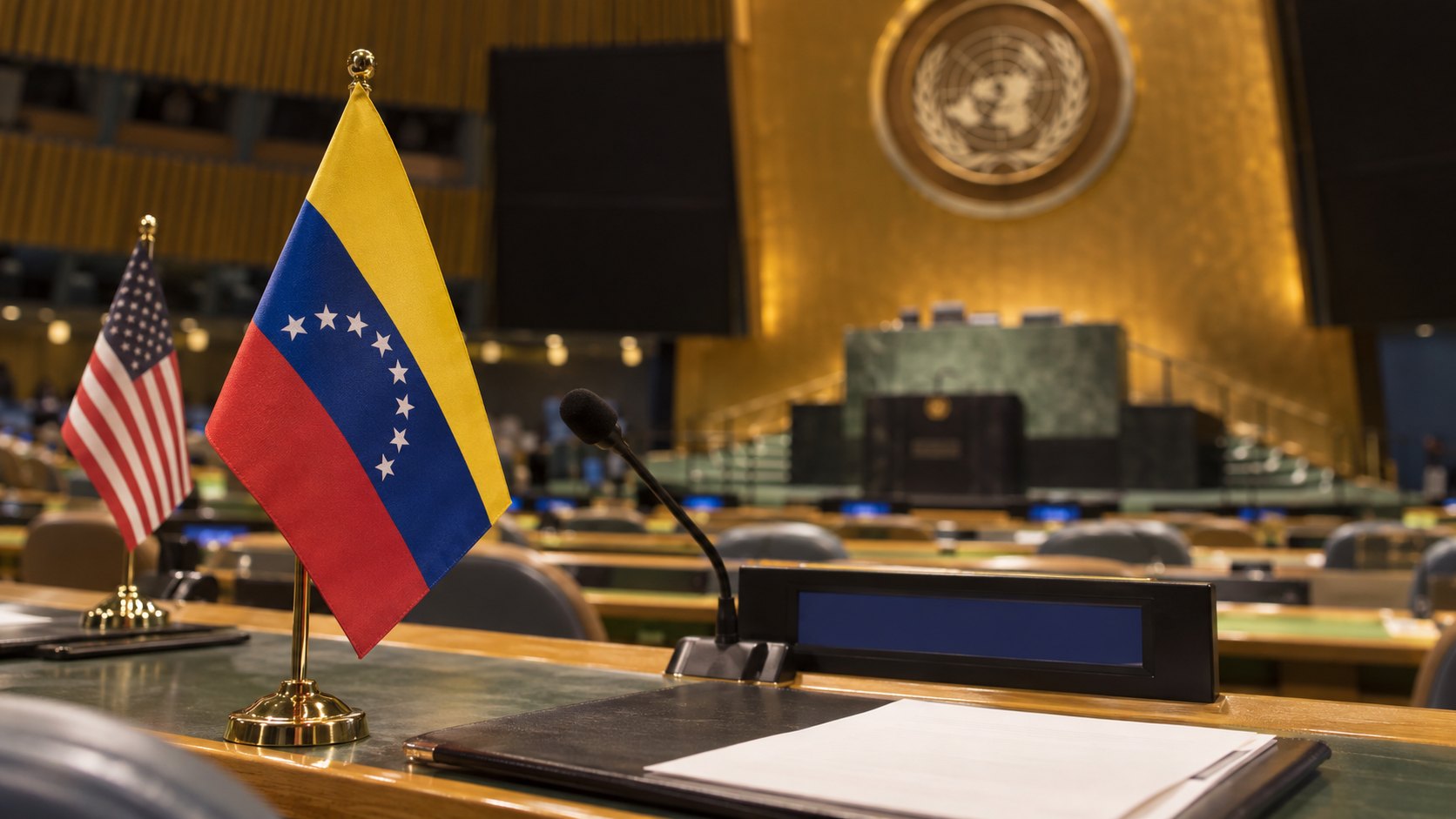 Ilustración editorial generada con IA: banderas de mesa de Venezuela y de Estados Unidos sobre un pupitre de delegación en la sala de la Asamblea General de la ONU, con el estrado y el emblema de Naciones Unidas al fondo; no es una fotografía del discurso.