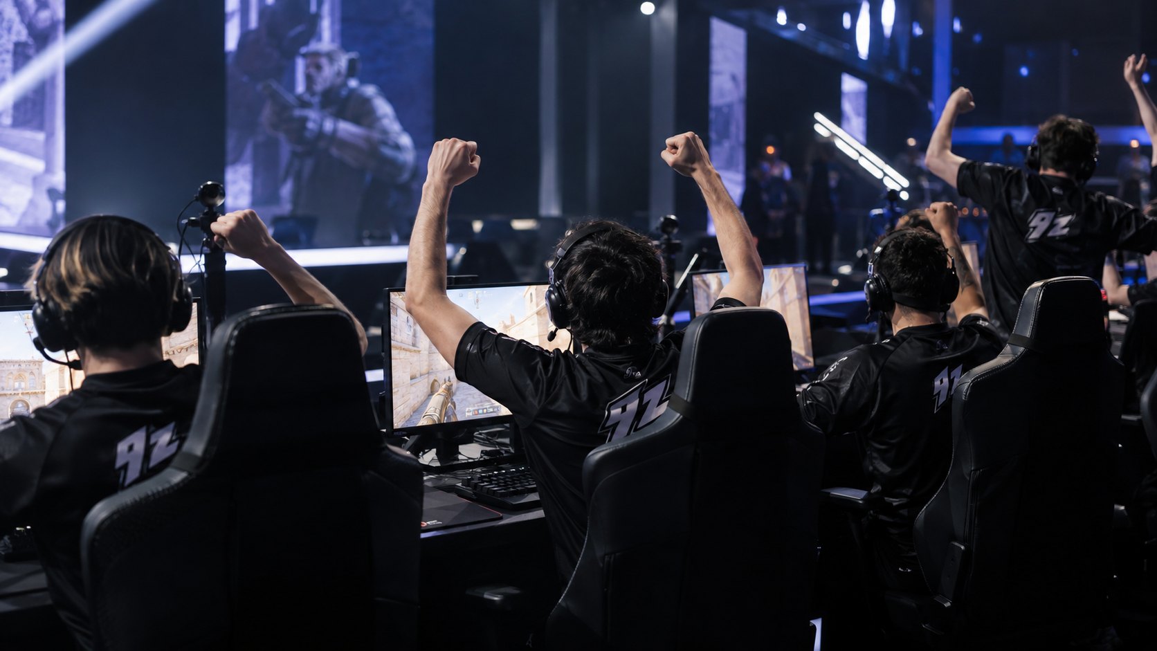 Ilustración editorial generada con IA: jugadores con camisetas negras con el logo de 9z, de espaldas, celebran con los brazos en alto en sus puestos de un estudio de e-sports, frente a monitores con Counter-Strike 2 y pantallas gigantes; composición conceptual, no es una fotografía del partido ni de los jugadores reales.