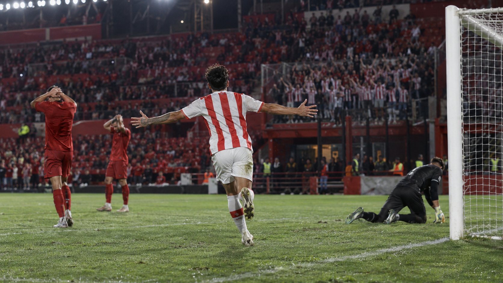 Ilustración editorial generada con IA: de noche, en un estadio de tribunas rojas, un futbolista de espaldas con camiseta blanca a rayas rojas festeja un gol con los brazos abiertos mientras dos jugadores con camiseta roja se lamentan junto al arco; no es una fotografía del partido ni representa a jugadores reales.