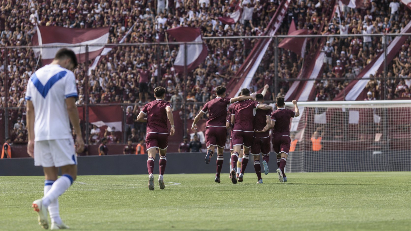 Ilustración editorial generada con IA: futbolistas con camiseta granate corren a festejar un gol frente a una tribuna llena de hinchas de Lanús, mientras un jugador con la camiseta blanca y la V azul de Vélez camina cabizbajo en primer plano; no es una fotografía del partido ni representa a jugadores reales.