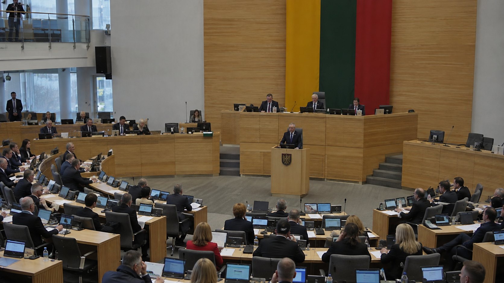 Ilustración editorial generada con IA: la sala de sesiones del Seimas de Lituania en Vilna, con legisladores en sus bancas y la bandera lituana detrás del estrado; composición conceptual, no es una fotografía de la votación.