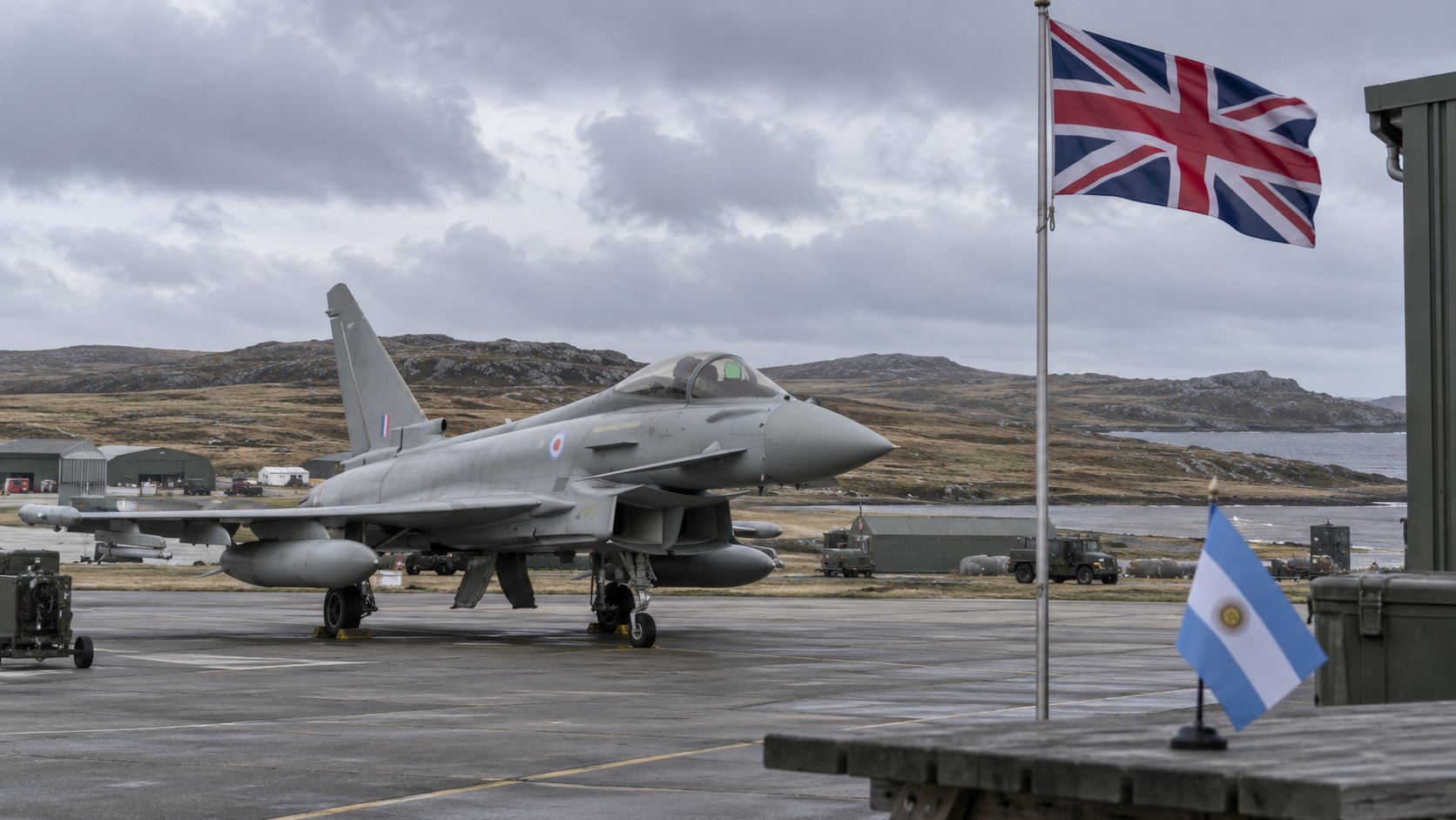 Ilustración editorial generada con IA: un caza Eurofighter Typhoon de la Real Fuerza Aérea británica en una pista bajo cielo gris, con una bandera del Reino Unido en un mástil, colinas de turba al fondo y una pequeña bandera argentina de mesa en primer plano; composición conceptual, no es una fotografía de la base de Mount Pleasant.