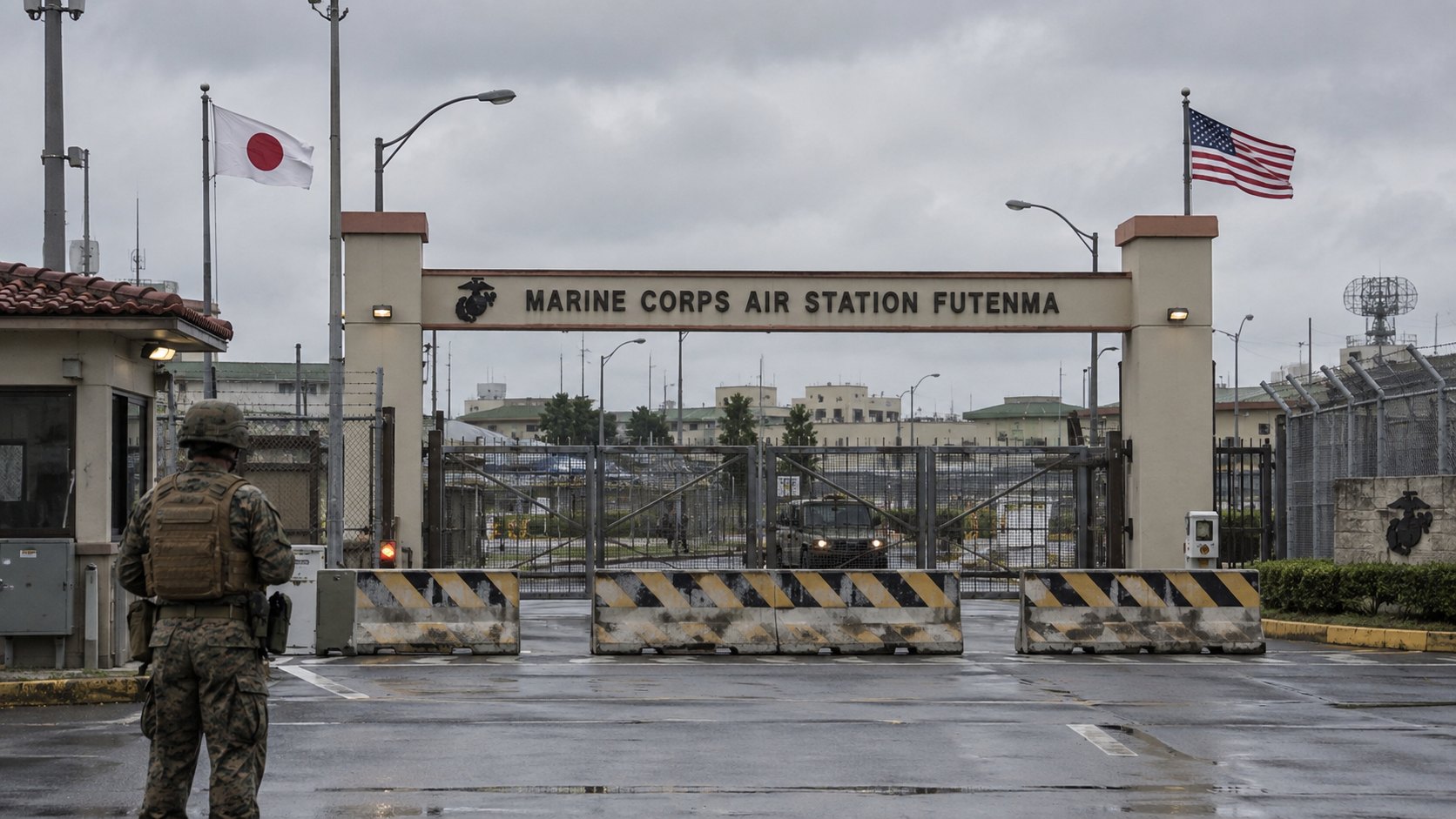 Ilustración editorial generada con IA: el portón de entrada de la Estación Aérea del Cuerpo de Marines de Futenma, en Okinawa, cerrado y custodiado por un guardia, con las banderas de Estados Unidos y de Japón en un día nublado; composición conceptual, no es una fotografía del lugar ni del hecho.