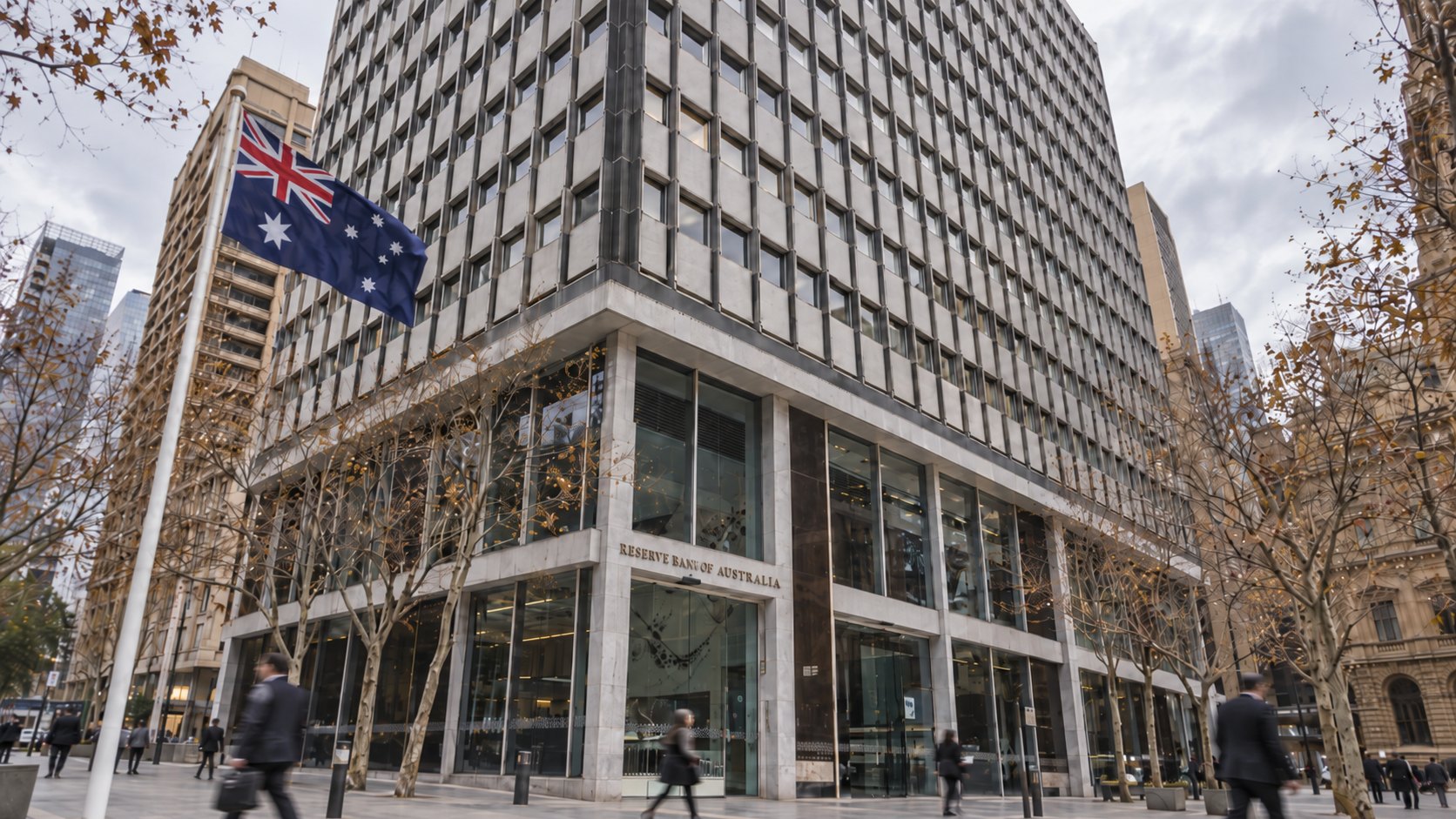 Ilustración editorial generada con IA: la sede del Reserve Bank of Australia en Martin Place, Sídney, con su nombre sobre la entrada y una bandera de Australia en primer plano, en una mañana nublada con peatones desenfocados; no es una fotografía del edificio ni de la reunión.
