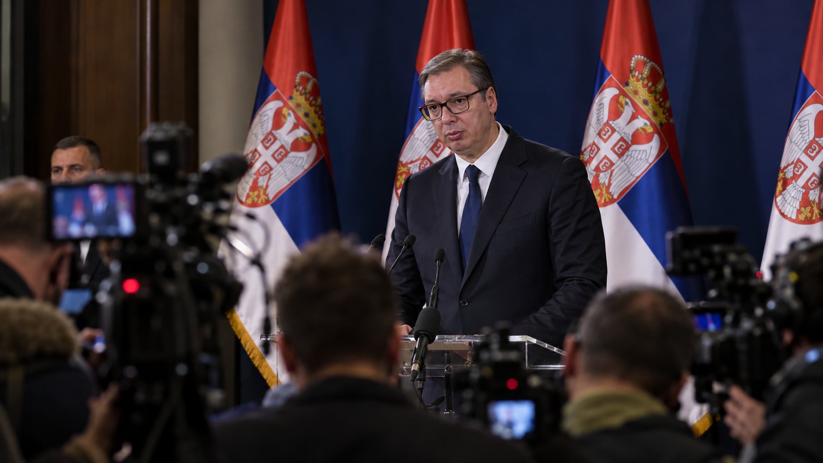 Ilustración editorial generada con IA: Aleksandar Vučić habla de pie ante un atril con micrófonos, frente a periodistas y cámaras de televisión, con banderas de Serbia detrás; composición conceptual, no es una fotografía de la conferencia del 25 de octubre.