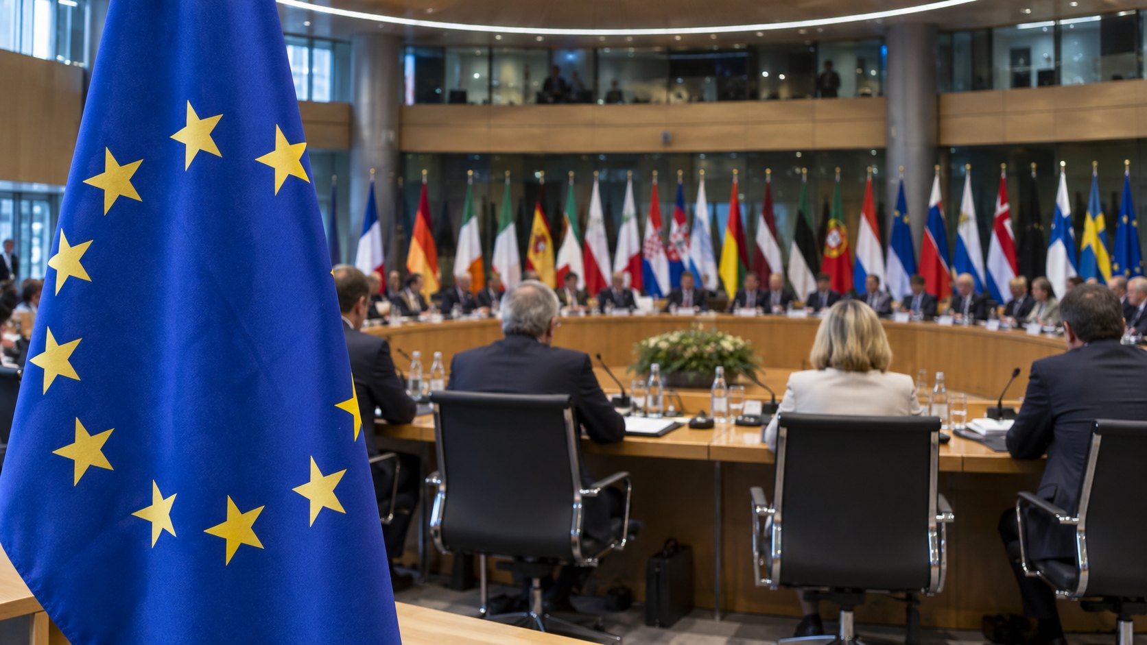 Ilustración editorial generada con IA: la sala de reuniones de los cancilleres de la Unión Europea en Luxemburgo, con la bandera de la UE en primer plano, banderas de los Estados miembros al fondo y ministros de espaldas alrededor de la mesa; composición conceptual, no es una fotografía del Consejo del 12 de octubre.