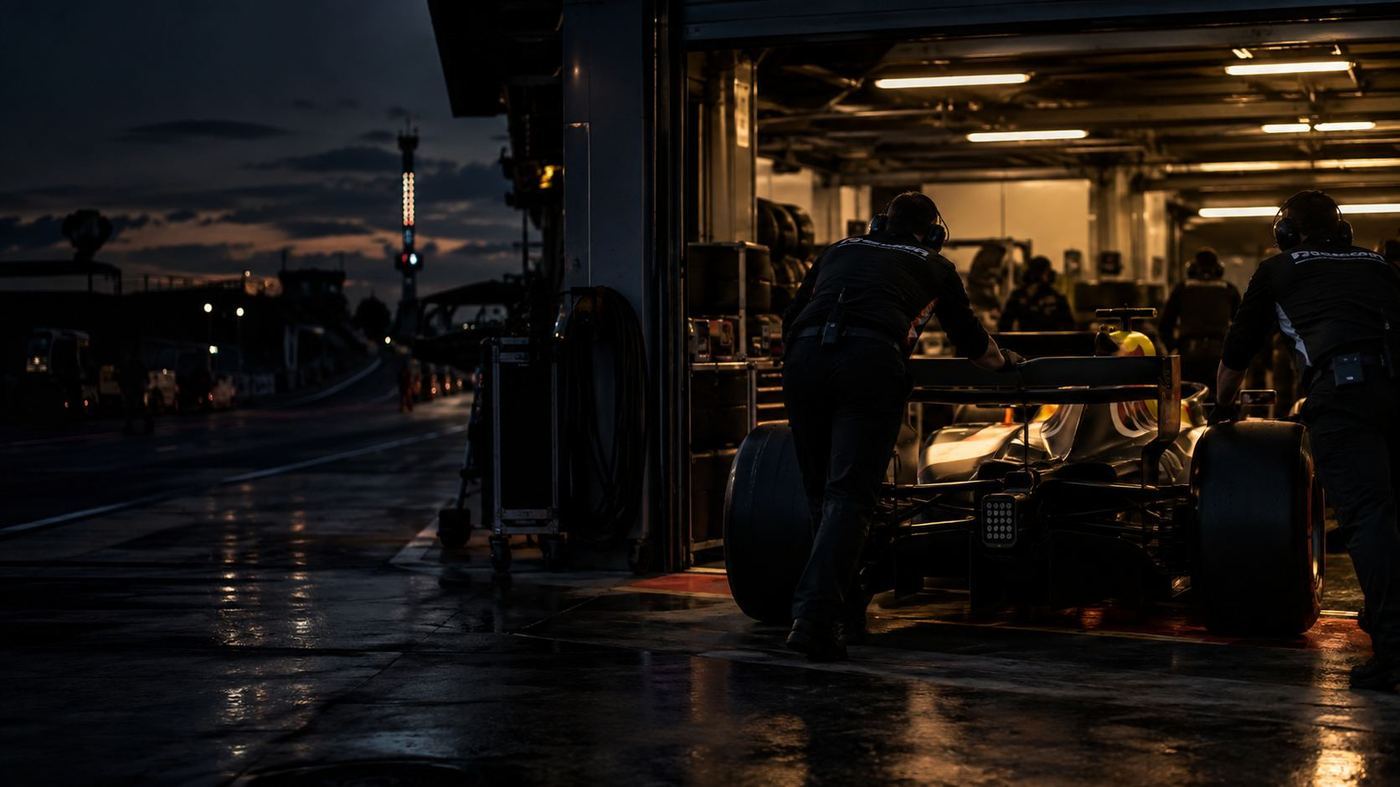 Monoplaza de Fórmula 2 siendo empujado hacia el box de un equipo en boxes, con mecánicos trabajando alrededor al atardecer