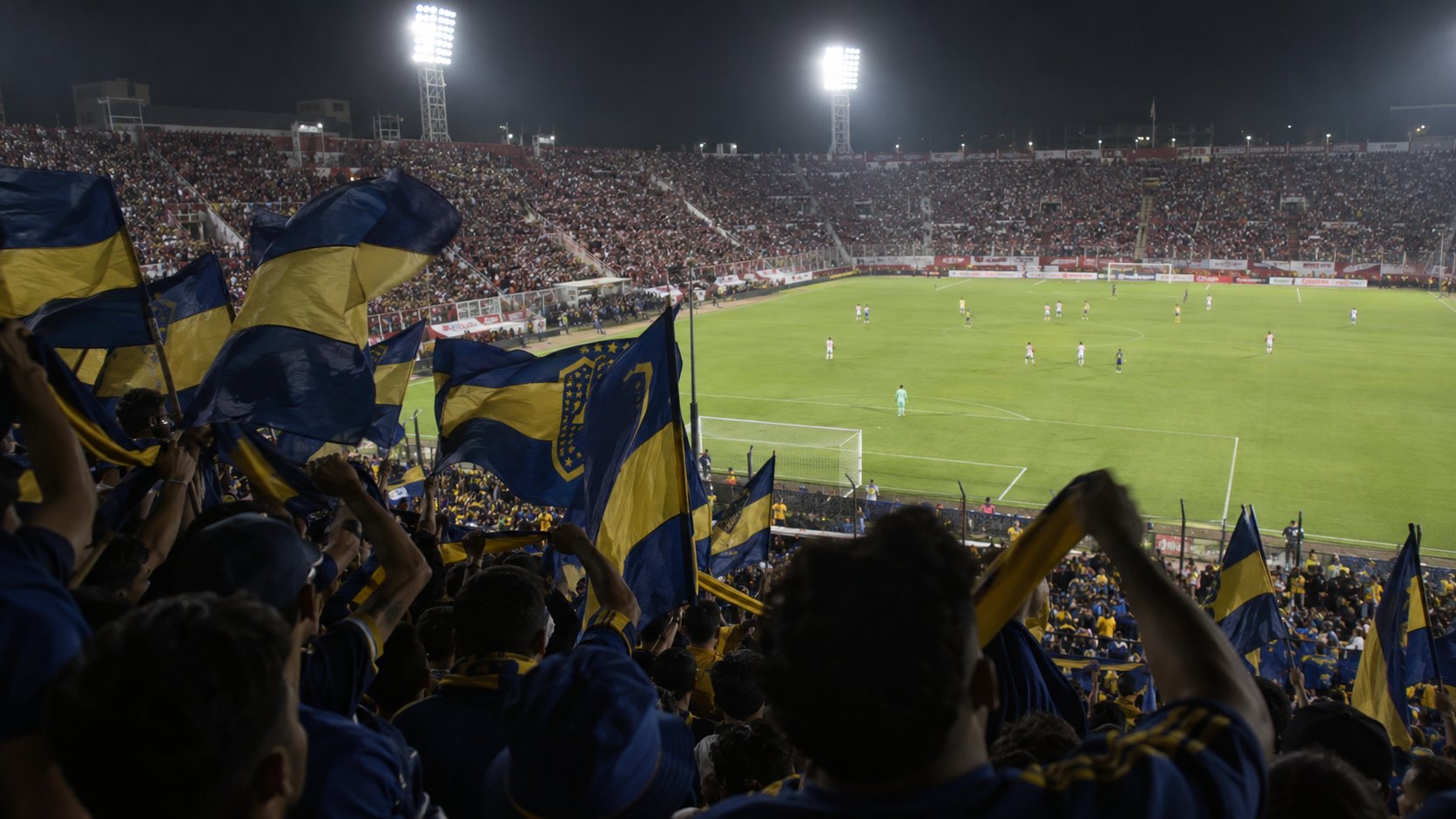 Ilustración editorial generada con IA: hinchas con banderas azul y oro de Boca Juniors agitan trapos en la tribuna de un estadio argentino iluminado de noche, con el campo y los jugadores a lo lejos; composición conceptual, no es una fotografía de un partido real.