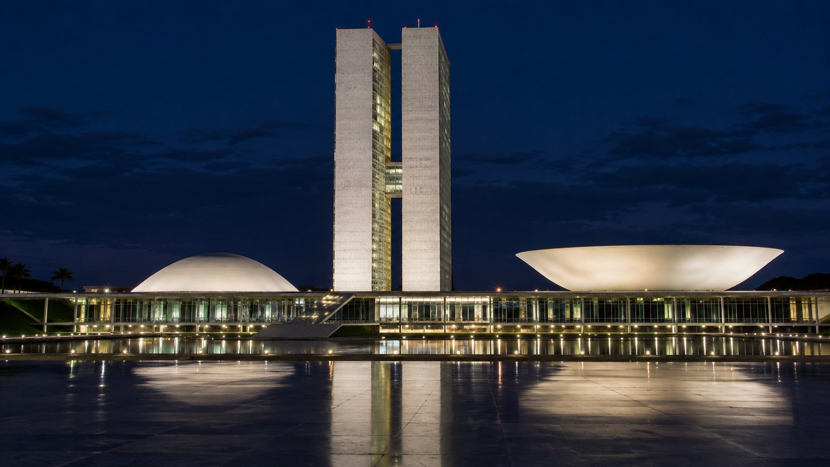 Ilustración editorial generada con IA: el edificio del Congreso Nacional de Brasil en Brasilia de noche, con sus dos torres y sus dos cúpulas iluminadas reflejadas en la explanada mojada; composición conceptual, no es una fotografía de la campaña.