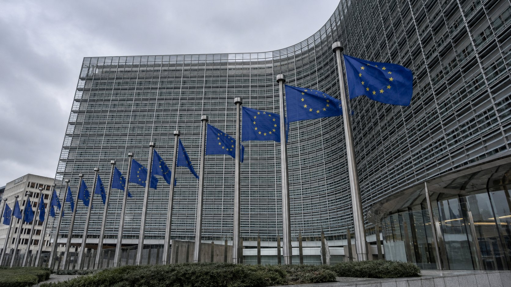 Ilustración editorial generada con IA: el edificio Berlaymont de la Comisión Europea en Bruselas con una fila de banderas de la Unión Europea al frente, en una mañana nublada; composición conceptual, no es una fotografía del día del anuncio.