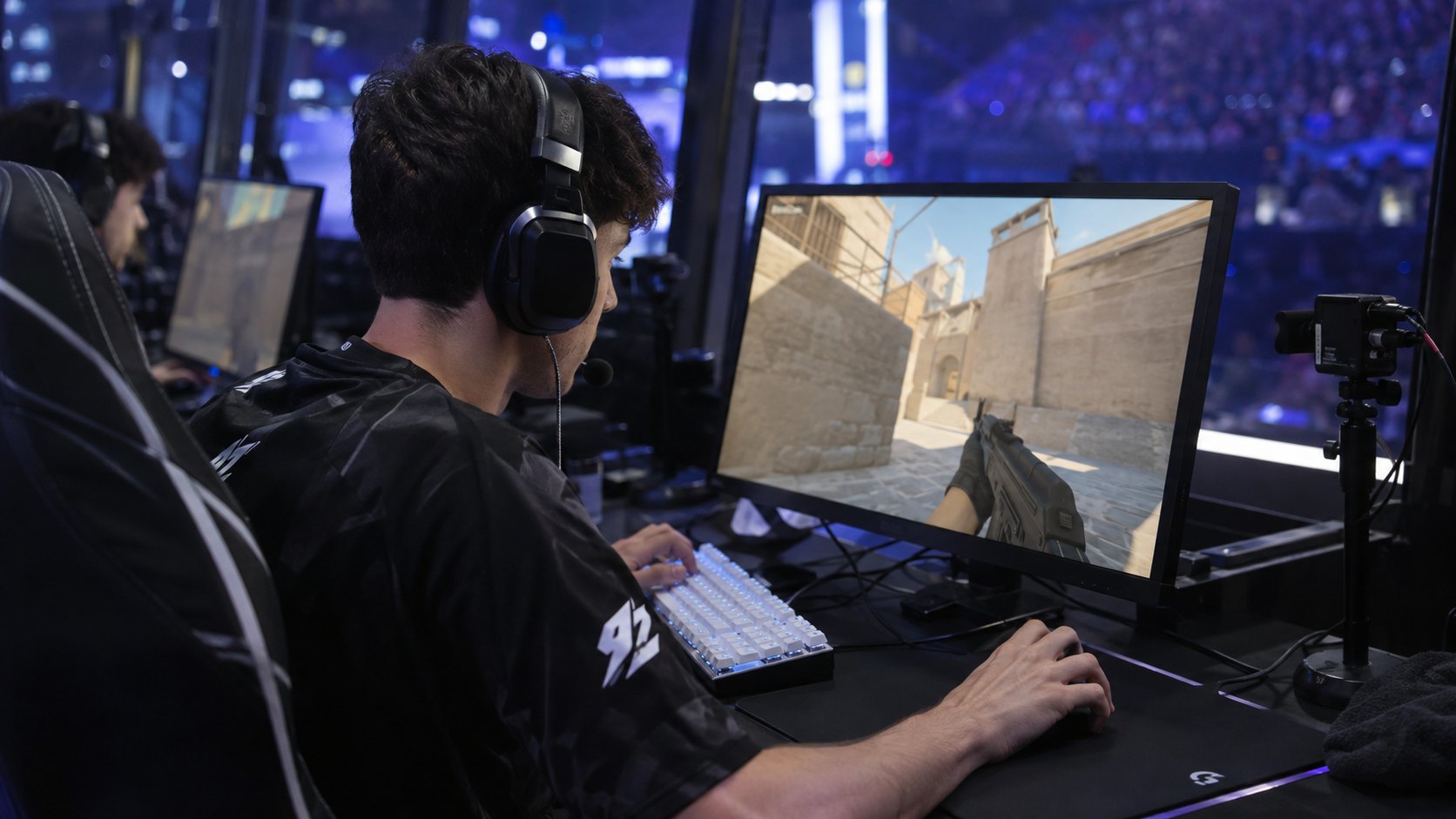 Ilustración editorial generada con IA: un jugador de 9z con auriculares, concentrado frente a su monitor durante una partida de Counter-Strike 2 en la cabina del escenario; no es una fotografía del partido.