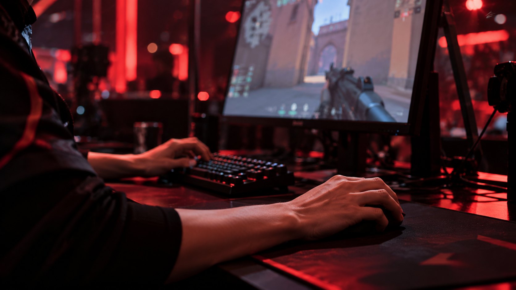 Ilustración editorial generada con IA: primer plano de las manos de un jugador sobre el teclado y el mouse en una cabina de torneo, con el monitor mostrando una partida de VALORANT y luces rojas del escenario; composición conceptual, no es una fotografía de un jugador real.