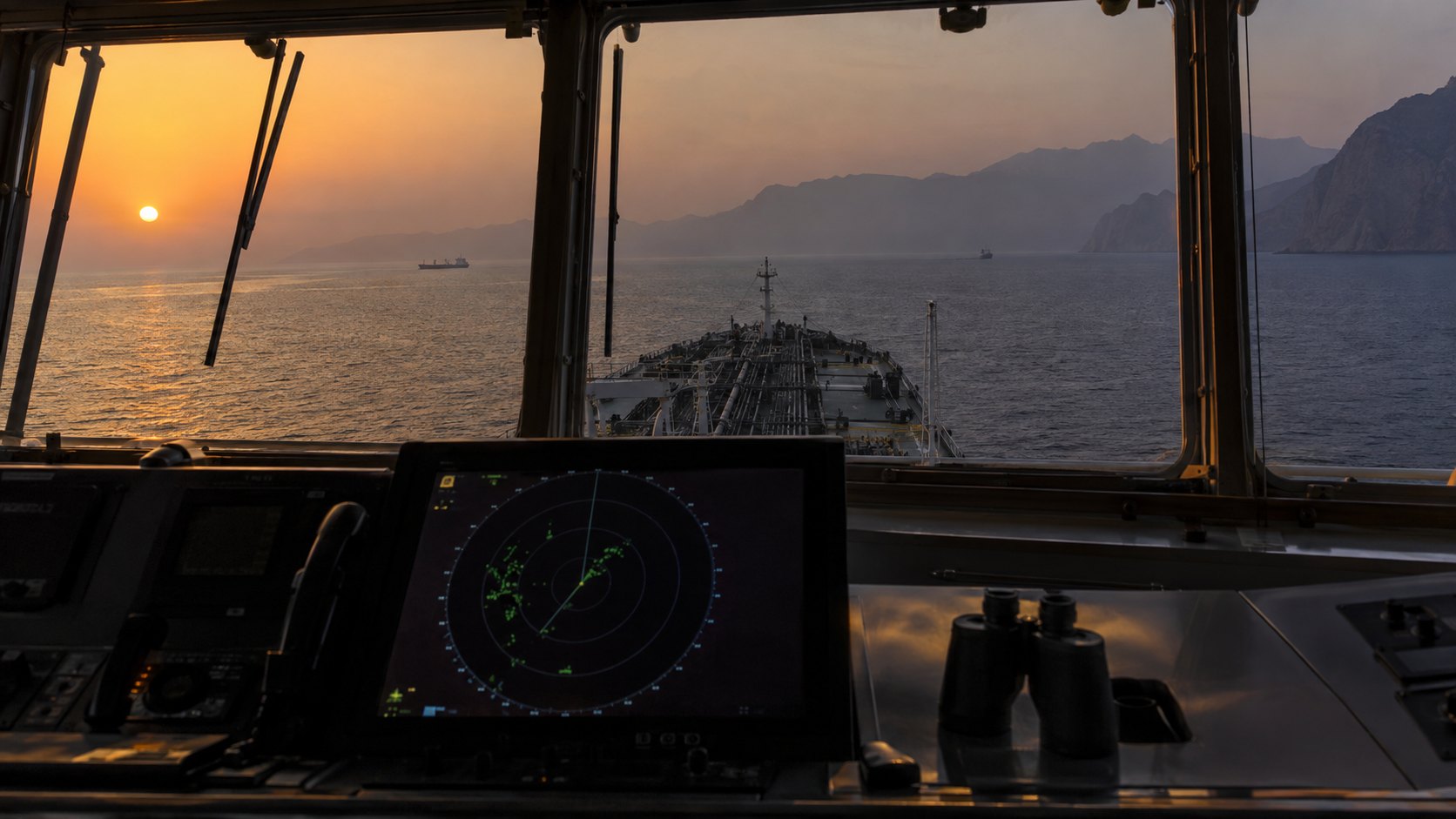 Ilustración editorial generada con IA: vista desde el puente de mando de un buque mercante que cruza el estrecho de Ormuz al atardecer, con un radar encendido y binoculares sobre la consola, pocas embarcaciones en el agua y las montañas de Musandam en la bruma; composición conceptual, no es una fotografía de un buque real.