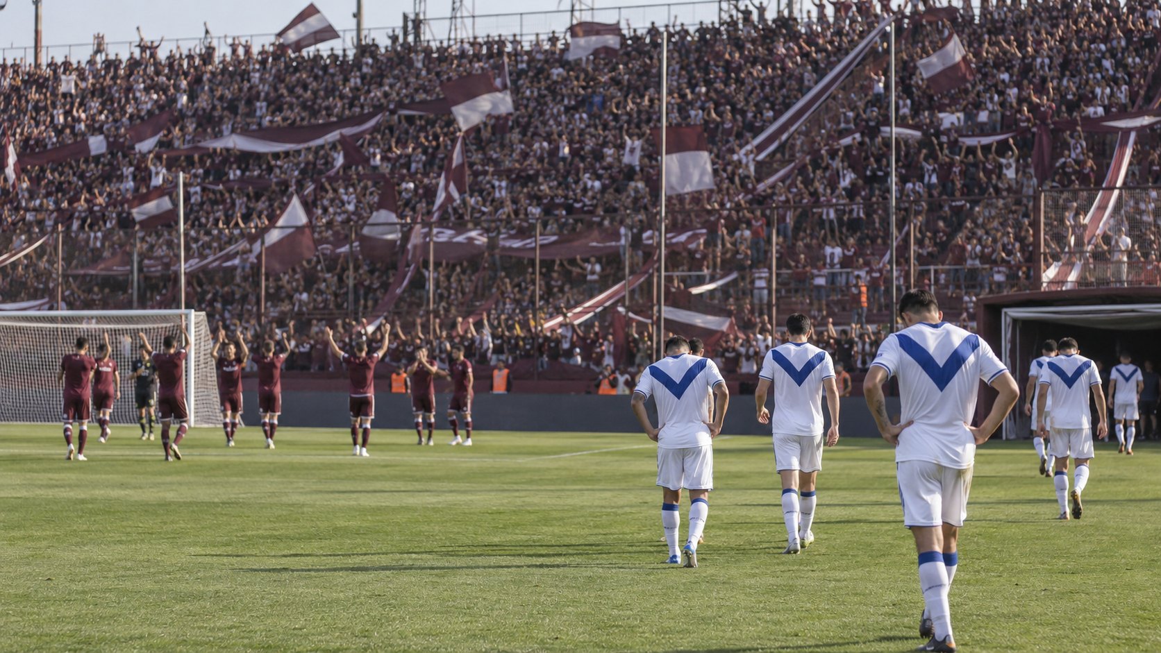 Ilustración editorial generada con IA: al final del partido, futbolistas de Vélez con camiseta blanca y V azul caminan de espaldas hacia el vestuario mientras los de Lanús, de granate, saludan a su hinchada en una tribuna llena de banderas granate y blancas; no es una fotografía del partido ni representa a jugadores reales.