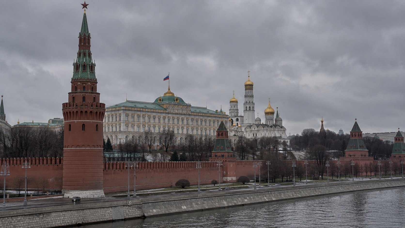 Ilustración editorial generada con IA: las murallas y torres del Kremlin de Moscú junto al río, con la bandera rusa sobre el Gran Palacio en un día nublado; composición conceptual, no es una fotografía del día de la declaración.