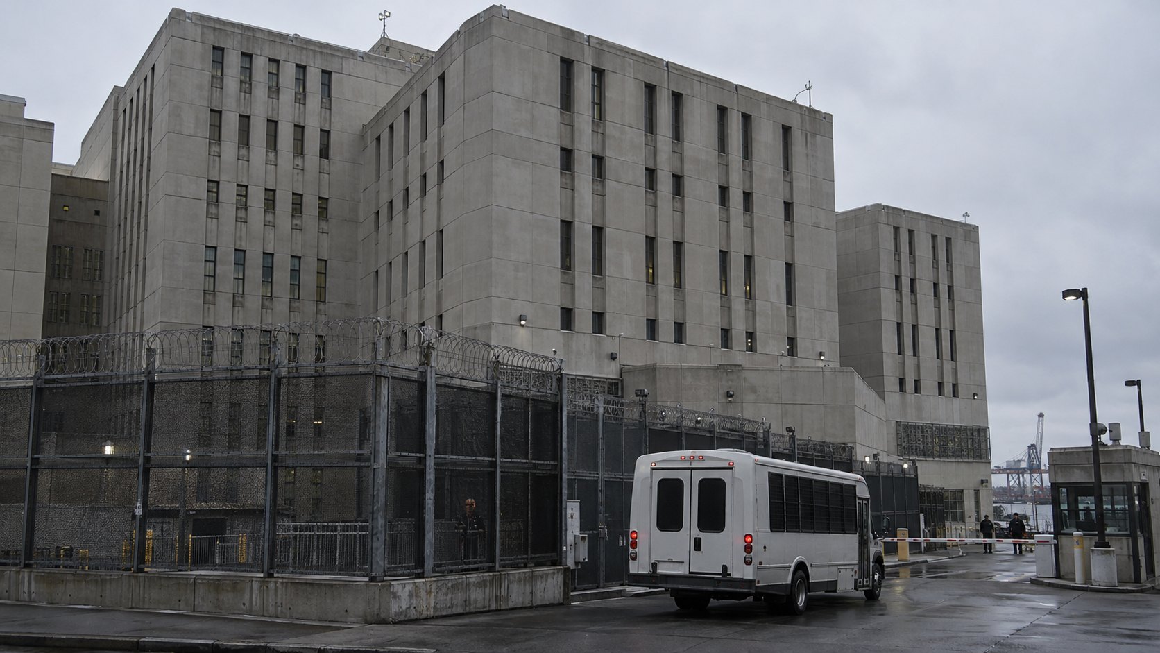Ilustración editorial generada con IA: el exterior del Metropolitan Detention Center de Brooklyn en un día nublado, con su cerco perimetral de alambre de púas y una camioneta blanca de traslado de presos en el acceso; composición conceptual, no es una fotografía de un traslado real.