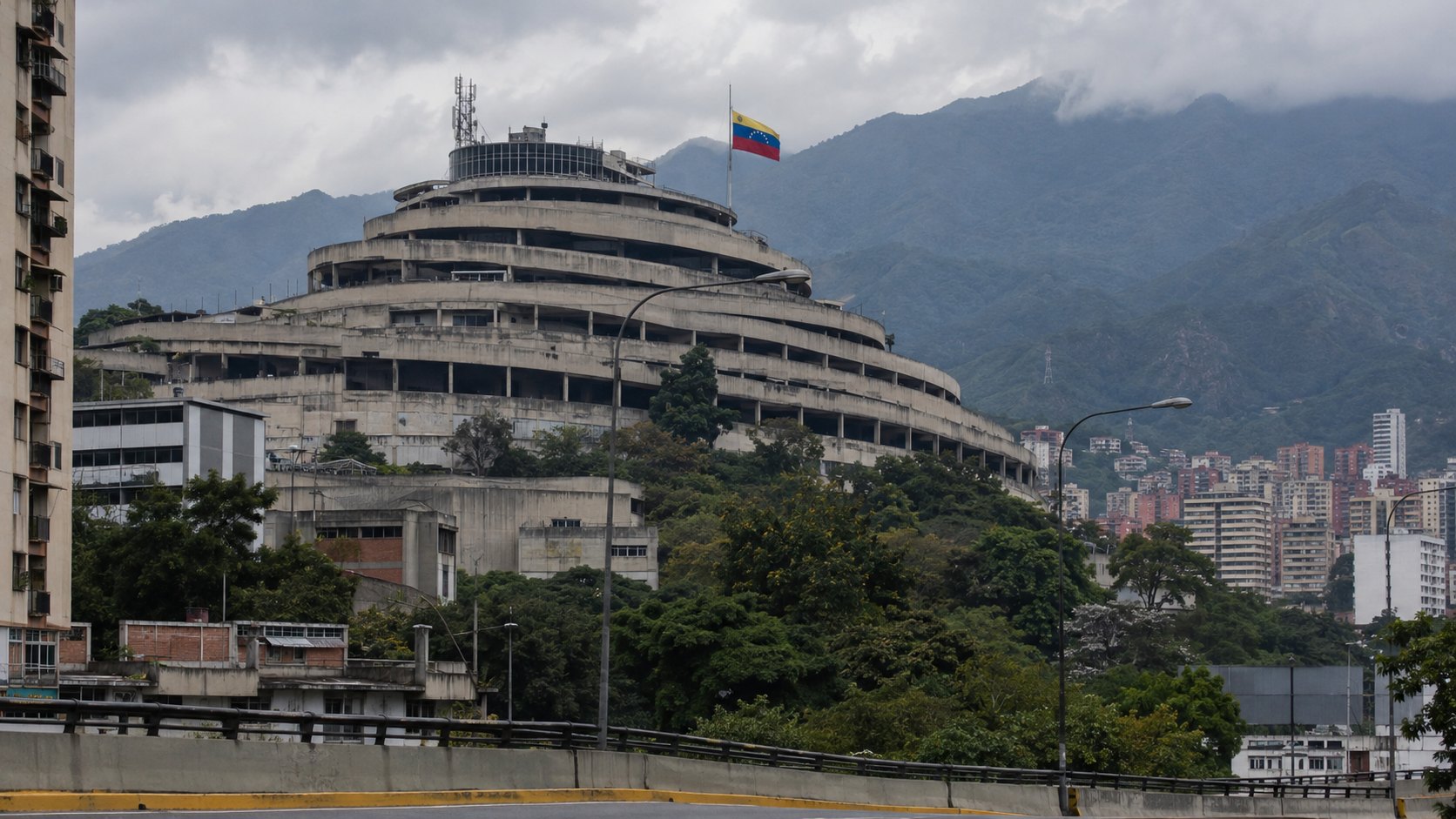 Ilustración editorial generada con IA: El Helicoide de Caracas, el edificio en espiral que el servicio de inteligencia venezolano usó como centro de detención durante el gobierno de Maduro, en una tarde nublada con el cerro Ávila al fondo y una bandera de Venezuela; composición conceptual, no es una fotografía documental.
