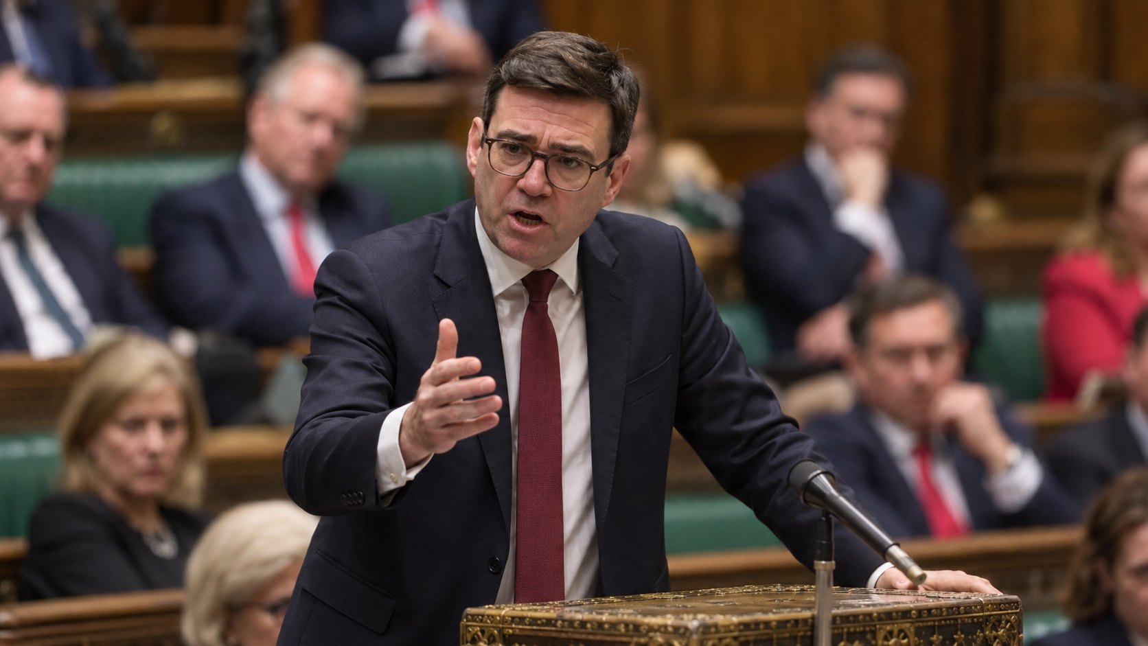 Ilustración editorial generada con IA: el primer ministro británico Andy Burnham habla en la tribuna del despacho de la Cámara de los Comunes, con diputados desenfocados en los bancos verdes detrás; composición conceptual, no es una fotografía de la sesión del 21 de octubre.