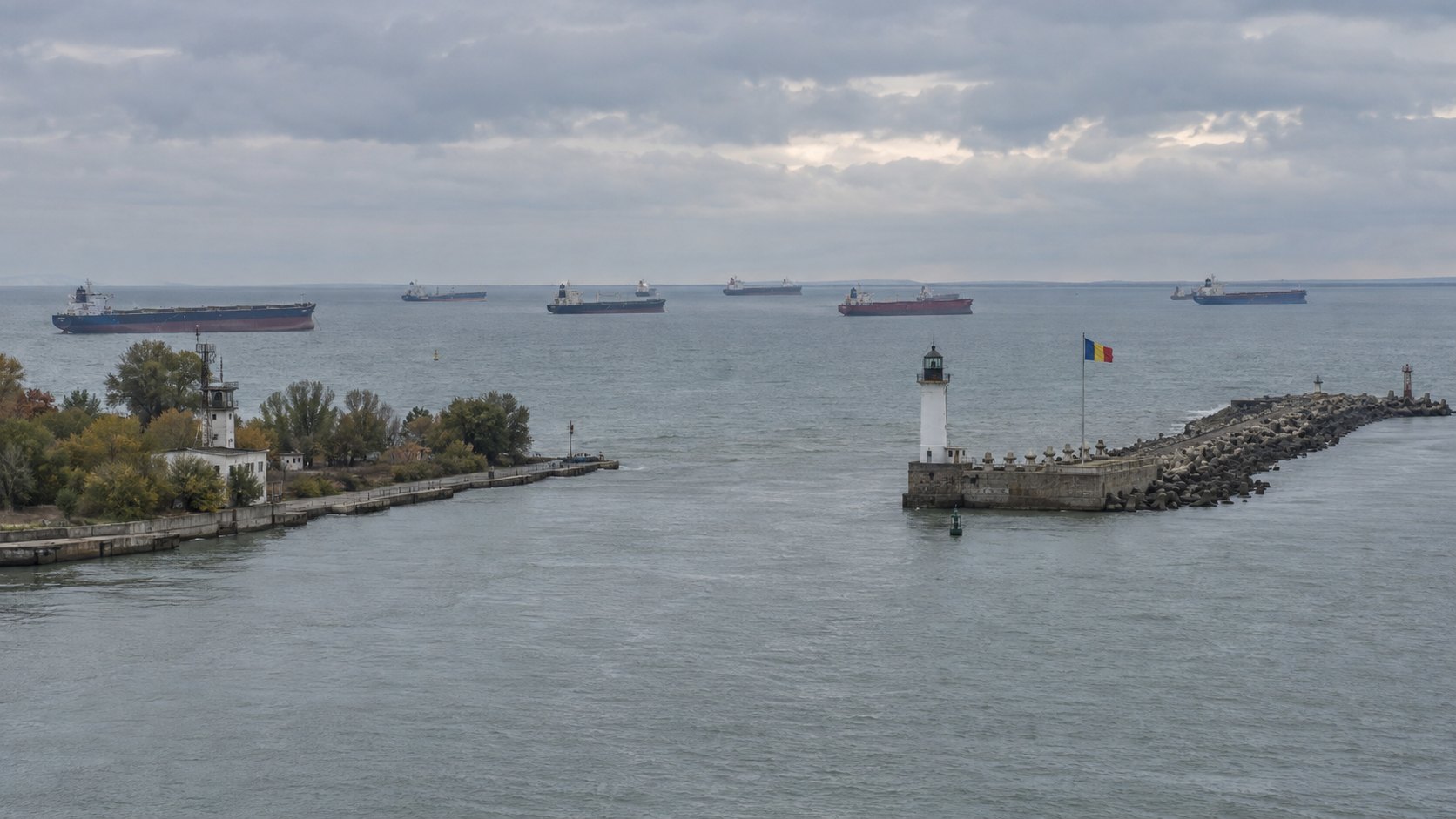 Ilustración editorial generada con IA: la desembocadura del canal de Sulina en el mar Negro, con el malecón, el faro y una bandera de Rumania, y buques graneleros y tanques fondeados a la espera de entrar al Danubio bajo un cielo nublado; composición conceptual, no es una fotografía del lugar ni del día.