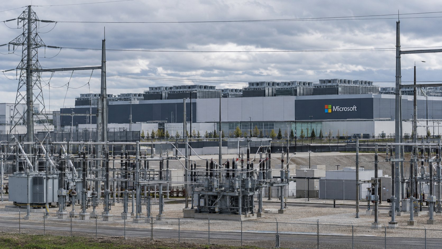 Ilustración editorial generada con IA: un campus de centros de datos con el logo de Microsoft en la fachada y una subestación eléctrica con líneas de alta tensión en primer plano; composición conceptual, no es una fotografía de una instalación real de Microsoft.