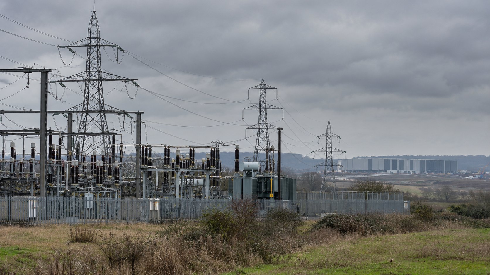 Ilustración editorial generada con IA: una subestación eléctrica de alta tensión vallada en la campiña inglesa, con torres de transmisión bajo un cielo nublado y, a lo lejos, el galpón gris de un centro de datos; composición conceptual, no es una fotografía de la red ni del sitio de Nscale.