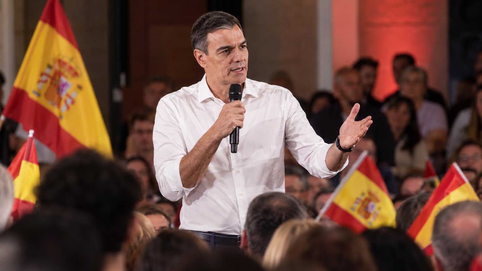 Ilustración editorial generada con IA: Pedro Sánchez, presidente del Gobierno de España, habla con un micrófono en la mano ante simpatizantes con banderas de España en un acto de campaña; composición conceptual, no es una fotografía del acto real en Burgos.