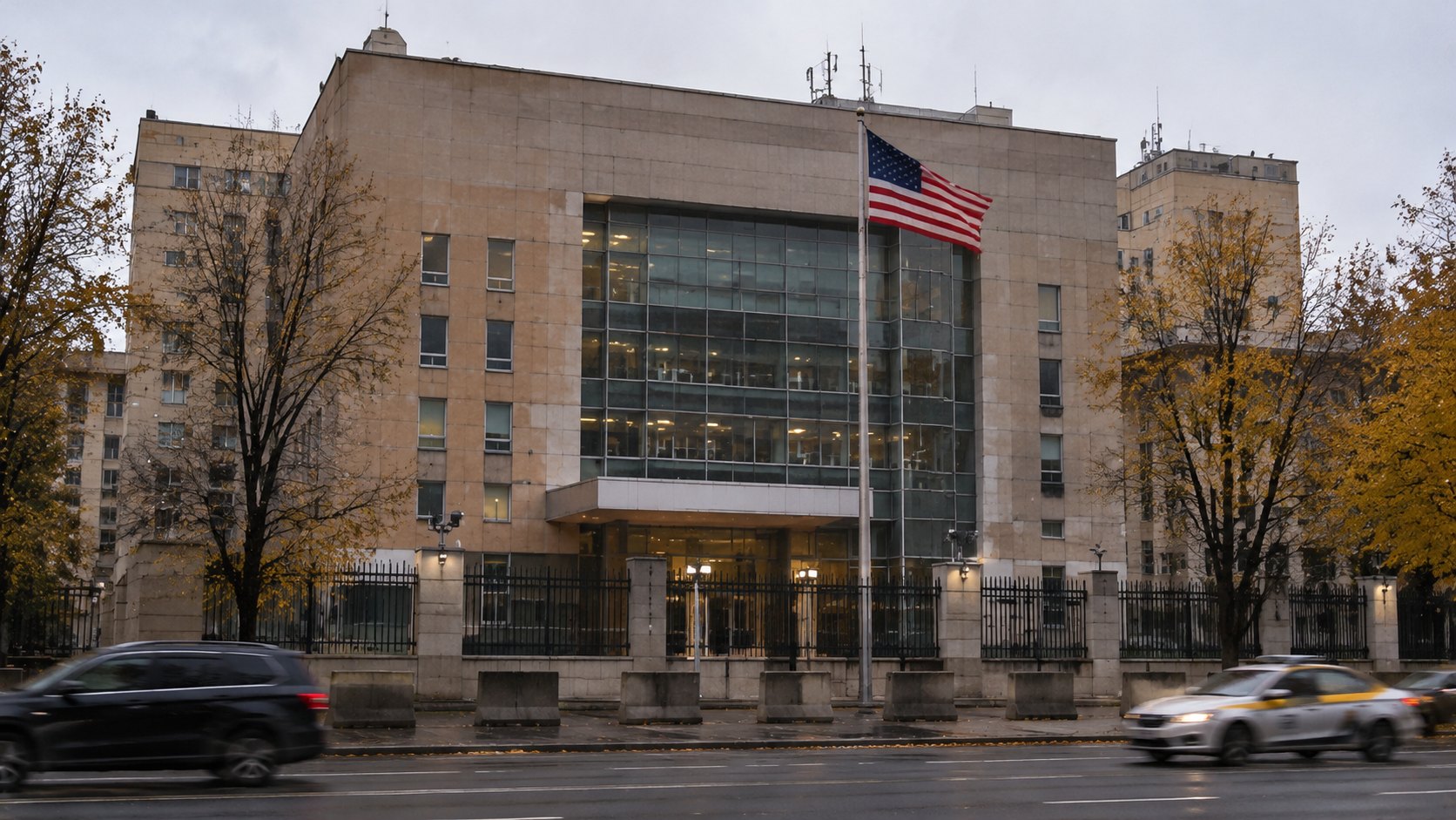 Ilustración editorial generada con IA: el edificio de la embajada de Estados Unidos en Moscú, con fachada de piedra clara y un frente vidriado, la bandera estadounidense en el mástil y tránsito en primer plano en una tarde gris de otoño; composición representativa, no es una fotografía del 31 de octubre.