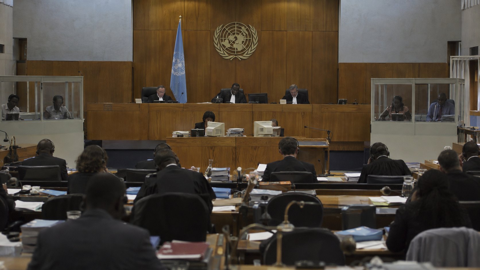 Ilustración editorial generada con IA: una sala de audiencias como la del Tribunal Penal Internacional para Ruanda, en Arusha, con el estrado bajo el emblema de las Naciones Unidas e intérpretes en sus cabinas; composición conceptual, no es una fotografía del juicio real.