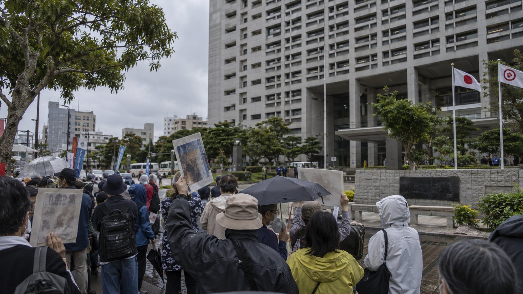 Ilustración editorial generada con IA: manifestantes con carteles y paraguas frente al edificio del gobierno de la prefectura de Okinawa, en Naha; composición conceptual, no es una fotografía de la protesta real.