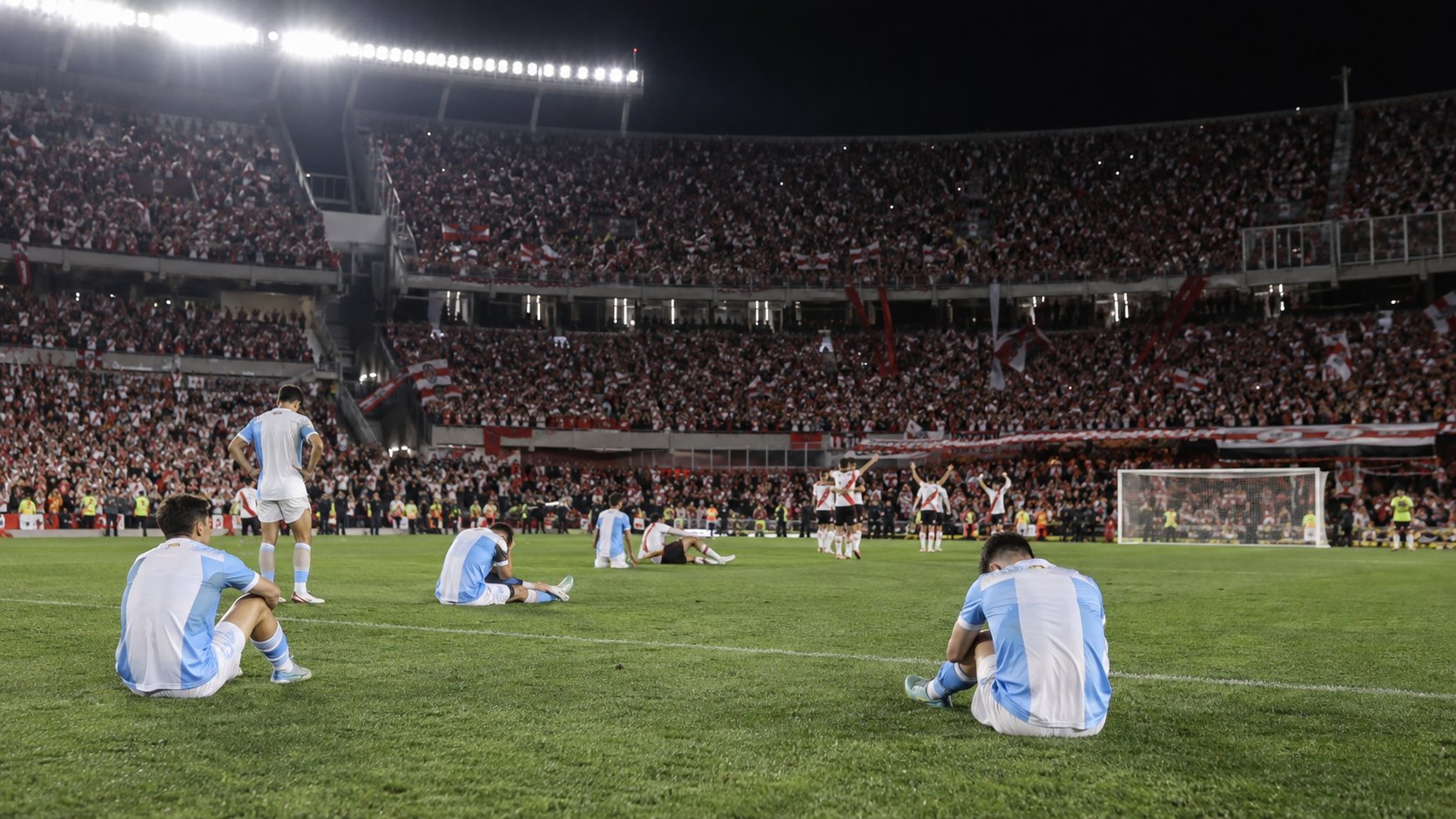 Ilustración editorial generada con IA: futbolistas visitantes sentados en el césped, cabizbajos y de espaldas, mientras la tribuna local festeja al fondo; no es una fotografía del partido ni representa a jugadores reales.